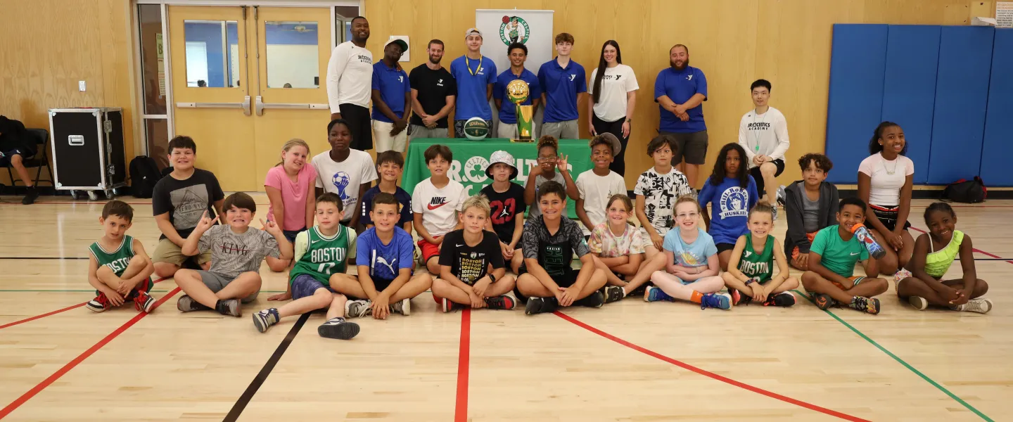 Group of kids from Basketball Camp sitting in front of the Celtics 2008 Championship trophy with YMCA coaches behind them