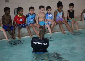 Kids sitting on the wall of a pool during their swim lesson with an instructor standing in front of them