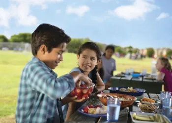 Boys and girls sitting at a picnic table outside eating food.